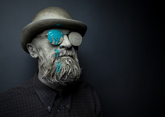 Portrait of a bearded man in with sunglasses. The face is covered with clay. On glasses green paint