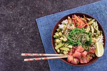 Homemade healthy Poké bowl with rice, salmon, avocado, white onion, scallion greens, sesame oil, soy and sesame sauce, seaweed (wakame) and other ingredients on a mat napkin and dark background