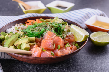 Homemade healthy Poké bowl with rice, salmon, avocado, white onion, scallion greens, sesame oil, soy and sesame sauce, seaweed (wakame) and other ingredients on a dark backgound close up