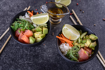 Two homemade healthy Poké bowl with rice, salmon, avocado, white onion, scallion greens, sesame oil, soy and sesame sauce, seaweed (wakame), lime and  rice vinegard on a dark backgound