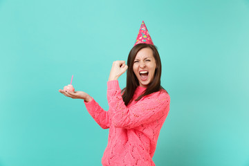 Happy young woman in knitted pink sweater, birthday hat screaming doing winner gesture, holding in hand cake with candle isolated on blue wall background. People lifestyle concept. Mock up copy space.