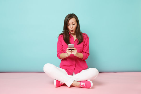 Full length portrait of young fun woman in rose shirt, white pants sit on floor hold cellphone isolated on bright pink blue pastel wall background studio. Fashion lifestyle concept. Mock up copy space