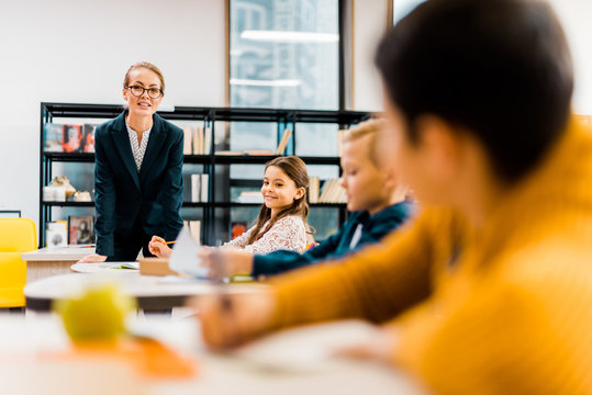 smiling young teacher looking at schoolchildren studying in library