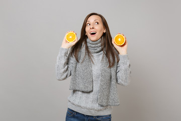 Curious young woman in gray sweater, scarf looking up and holding oranges isolated on grey wall background. Healthy fashion lifestyle, people sincere emotions, cold season concept. Mock up copy space.