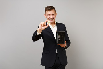 Cheerful young business man in suit holding bitcoin future currency, metal bank safe for money accumulation isolated on grey background. Achievement career wealth business concept. Mock up copy space.