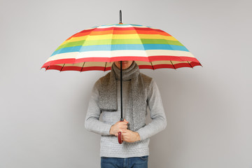 Young man in gray sweater, scarf covering face with colorful umbrella isolated on grey background...
