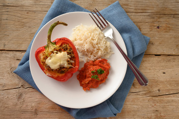 stuffed red bell pepper baked with cheese gratin, rice and tomato sauce on a plate and a blue napkin, rustic wooden table, copy space, high angle view from above