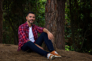 Naklejka na ściany i meble Handsome man on lumberjack check shirt sitting by pine tree in forest. Young hipster posing with urban clothing in nature. Fashion style model concept