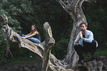 Young couple performing yoga asanas on tree trunk in forest. Man in malasana and woman in paschimottanasana pose in nature