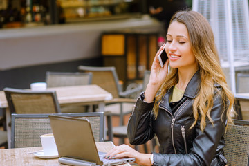 Attractive young woman smiling and talking on smartphone while sitting at table in outdoor cafe and browsing modern laptop