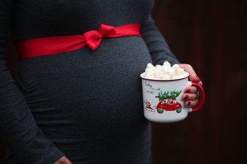 Pregnancy, Christmas and People Concept - close up of Pregnant Woman with Red ribbon over, holding Cup with Hot Coco and  Mushmellow 