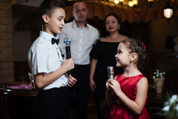 Brother and sister sing karaoke songs in microphones, and their parents sing in the back