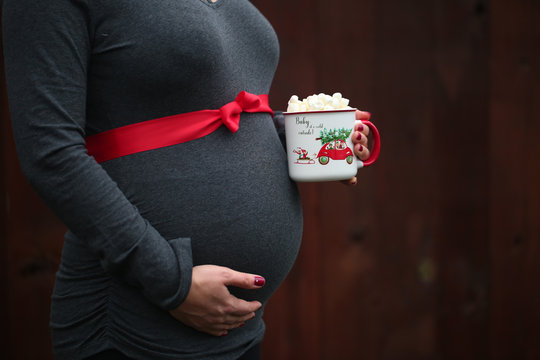 Pregnancy, Christmas and People Concept - close up of Pregnant Woman with Red ribbon over, holding Cup with Hot Coco and  Mushmellow 