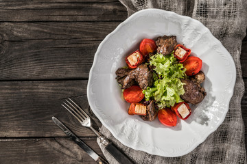 Hot salad with chicken liver, mushrooms, salad leaves, cheese, tomatoes on plate over rustic wooden background. Healthy food, top view