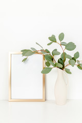 Blank frame mockup with a branches of eucalyptus in vase on table on light background. Home decor. Blog, website or social media concept.