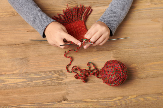 Child girl is learning to knit a scarf. Red wool yarn is on the wooden table. Hand closeup.