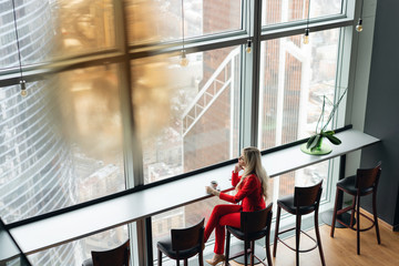 Woman in red coral business suit drinks coffee at a high table near the window. Young blonde businesswoman in the office in high floor in front of the window.