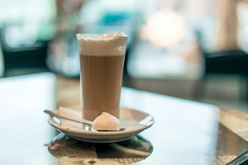 A cup of hot latte coffee in transparent glass on white plate with a sweet treat and teaspoon a glass table with light bokeh background, front view.