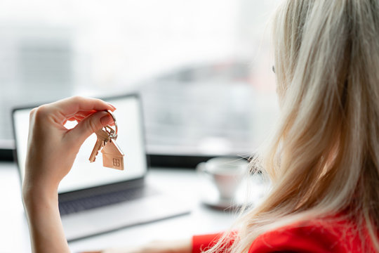 Mortgage concept. Woman in red coral business suit holding key with house shaped keychain. Modern light lobby interior. Real estate, hypothec, moving home or renting property. - Powered by Adobe