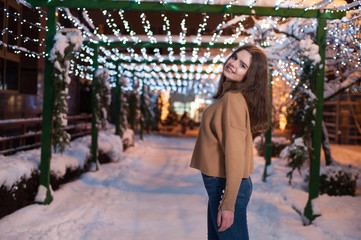 Pretty dark haired girl wearing blue jeans and beige top with snowflakes Christmas lights outdoor at night time.