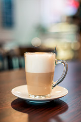 A cup of hot latte coffee in transparent glass on a wooden dark brown table and teaspoon in a cup with light bokeh background, front view.