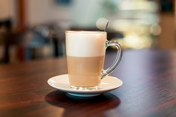 A cup of hot latte coffee in transparent glass on a wooden dark brown table and teaspoon in a cup with light bokeh background, front view.