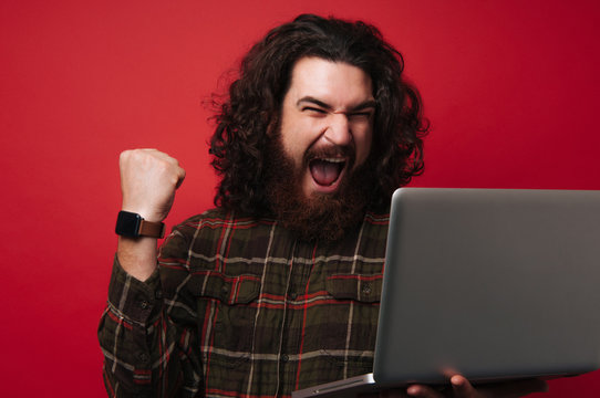 Winner bearded man with long hair, wearing shirtand looking at laptop screen, making a winner gesture