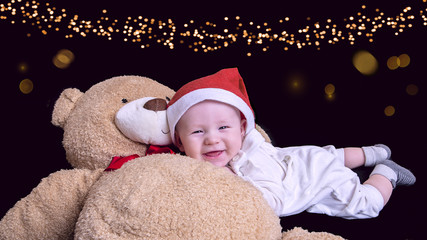 Christmas portrait of cute little newborn baby boy, wearing santa hat and hugging big cute bear toy, studio shot, winter time, black background with lens flare 