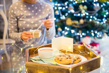 Christmas and New Year composition. Cocoa and cookies on the wooden tray and blurred no face woman opening her present on xmas background. Sweets for Santa. Festive breakfast. Soft selective focus.