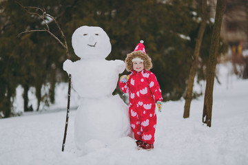Cute little girl in winter warm clothes with hood playing, standing near snowman in park or forest...
