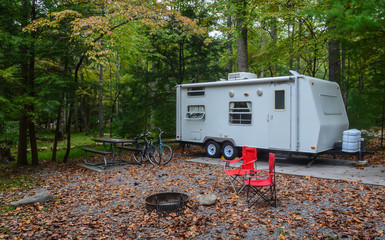 Camping in camper trailer in wooded park camp site with chairs set up around fire pit, and bicycles...