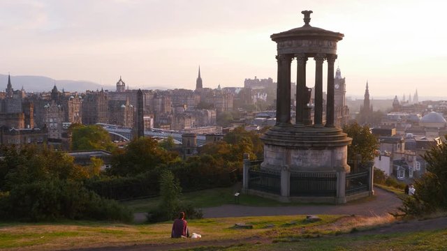View over Edinburgh from the Calton Hill with the Dugald Stewart Monument in the foreground and a girl laying on grass enjoying the view at sunset