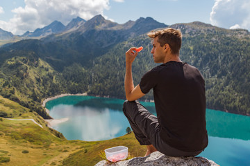 young man seated on a rock in the mountains eat watermelon and look to the panorama