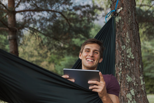 young man smiling to camera and using a tablet on a hammock