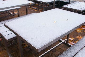 Snow-covered tables and benches in the Park in autumn with yellow leaves.
