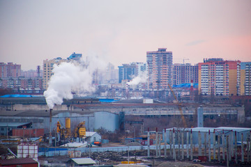 Landscape of evening city with high-rise buildings under construction, factories and pipes with smoke at sunset under boundless cloudy sky