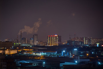 Landscape of night city with high-rise buildings under construction, factories and pipes with smoke under boundless cloudy sky