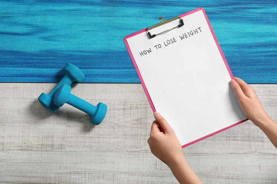 Woman holding clipboard with words HOW TO LOSE WEIGHT over sport equipment, top view
