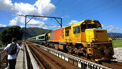 Train passing over old riverside bridge on a sunny New Zealand day with the hilly backdrop as a student crosses the bridge