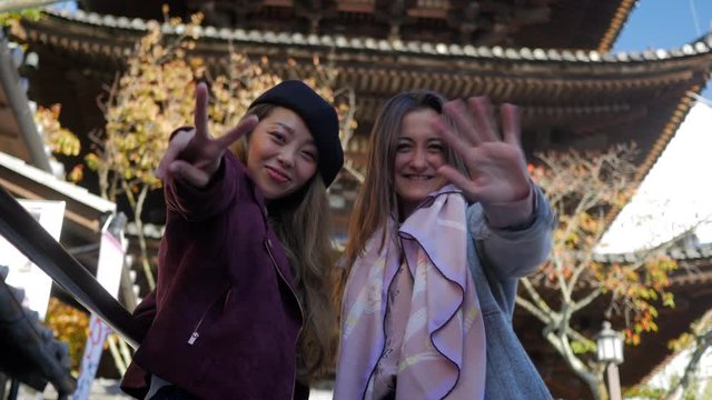 4K excited international women waving and smiling to camera, large Kyoto temple behind, low angle.