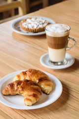 capuchino con dos croissants, medialunas y postre, torta, cake  sobre mesa de madera con luz natural