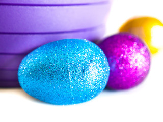 Close up of a sparkling blue, pink and yellow Easter egg sitting on a white table and purple plastic bucket or basket in the background making a perfect holiday backdrop.