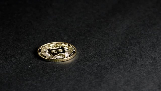 Golden Bitcoin spinning on a black surface. The minted coin comes in from the right, then spins through the picture and comes to rest on the left. The coin is polished and especially shiny.