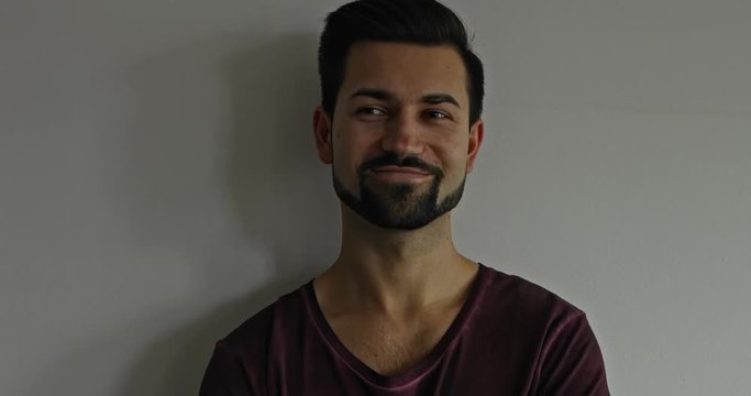 Handsome adult male leans against a while wall, his expression changing from serious to smiling