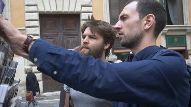 Two Young Male Tourists Trying To Choose A Postcard To Send Home.