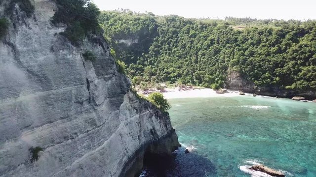 Aerial view of the rocks in Nusa Penida. Flying over white sand Atuh Baech, Indonesia. Tourists swimming in the ocean. Wonderful beach with umbrellas and lounge beds.