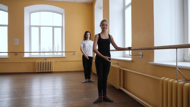 A beautiful ballerina dancer teaching a model friend how to perform the basic positions using a ballet bar in a studio with large bright windows