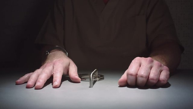 Close-up parallax shot of a prisoner's hands as he sits handcuffed to a table in an interrogation room.