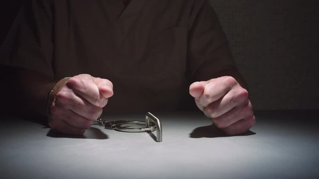 Close-up parallax shot of a prisoner's hands as he sits handcuffed to a table in an interrogation room, his hands balled into fists.