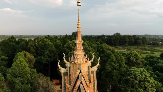 Aerial drone view orbiting the ornate roof and spire of a Khmer style Buddhist temple in Southeast Asia; as camera moves higher and circles, forest and river is revealed in background.